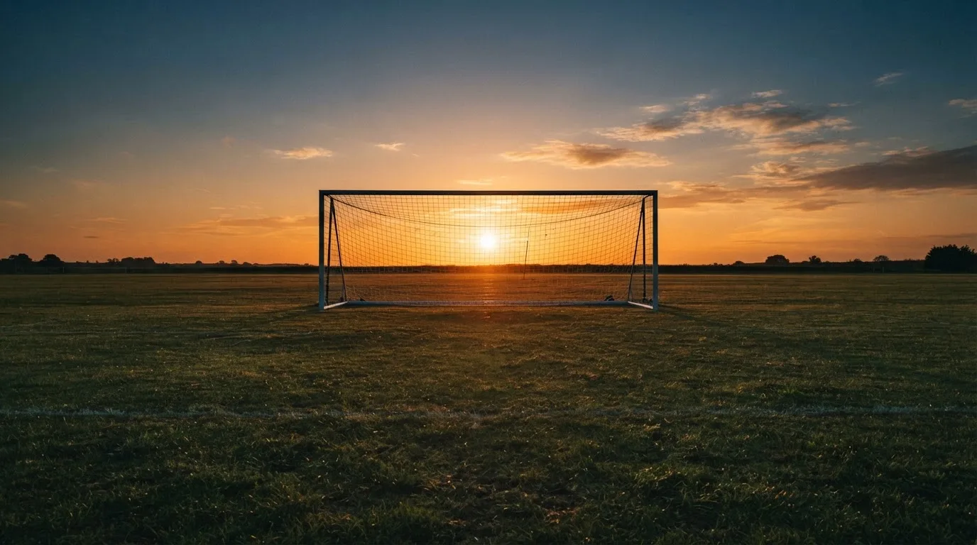 Ein Fußballtor mit Netz bei Sonnenuntergang auf einem leeren Platz, die Schatten der Torpfosten liegen lang auf dem Gras.