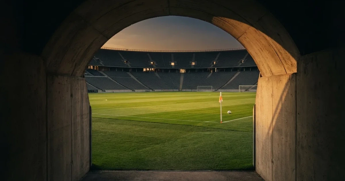 Blick durch ein Stadiontor auf einen grünen Rasen im Abendlicht, ein Ball liegt an der Eckfahne, die Ränge sind leer.