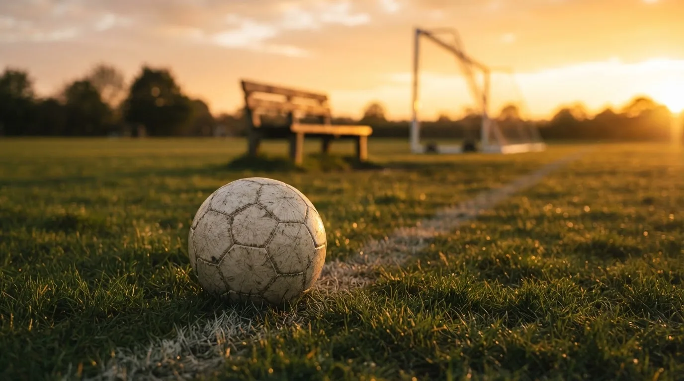 Ein Fußball liegt im Gras am Spielfeldrand, dahinter eine leere Bank und ein Tor im Gegenlicht.