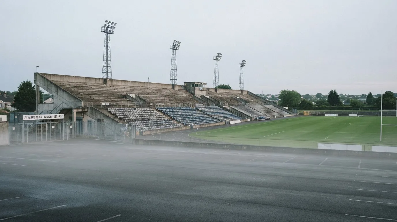 Eine leere Tribüne eines kleinen Stadions am frühen Morgen, aus der Ferne über einen menschenleeren Parkplatz fotografiert.