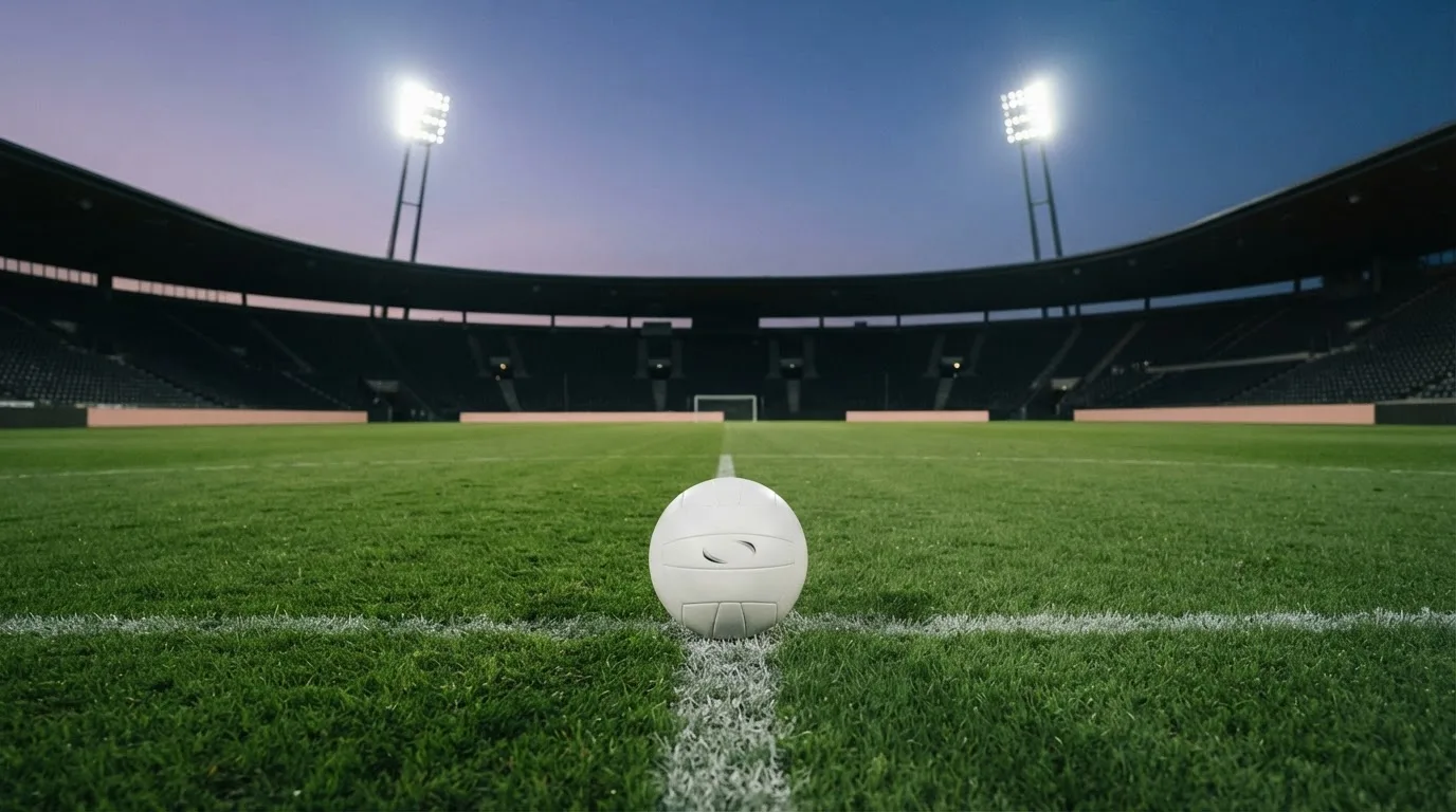 Ein Fußball liegt auf dem Rasen eines leeren Platzes, im Hintergrund ein Stadion unter Flutlicht in der Abenddämmerung.