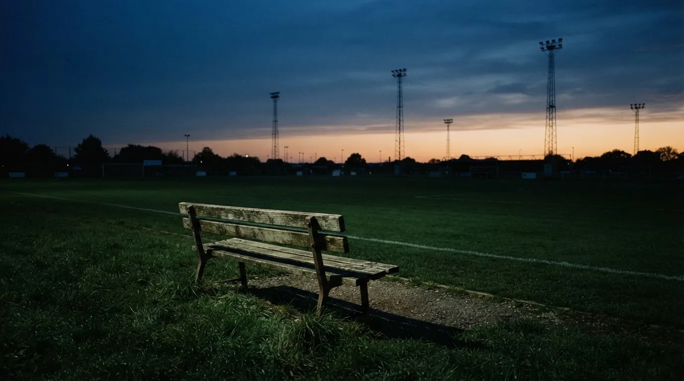 Eine leere Parkbank am Rand eines Fußballfelds in der Abenddämmerung, im Hintergrund ragen dunkle Flutlichtmasten in den Himmel.