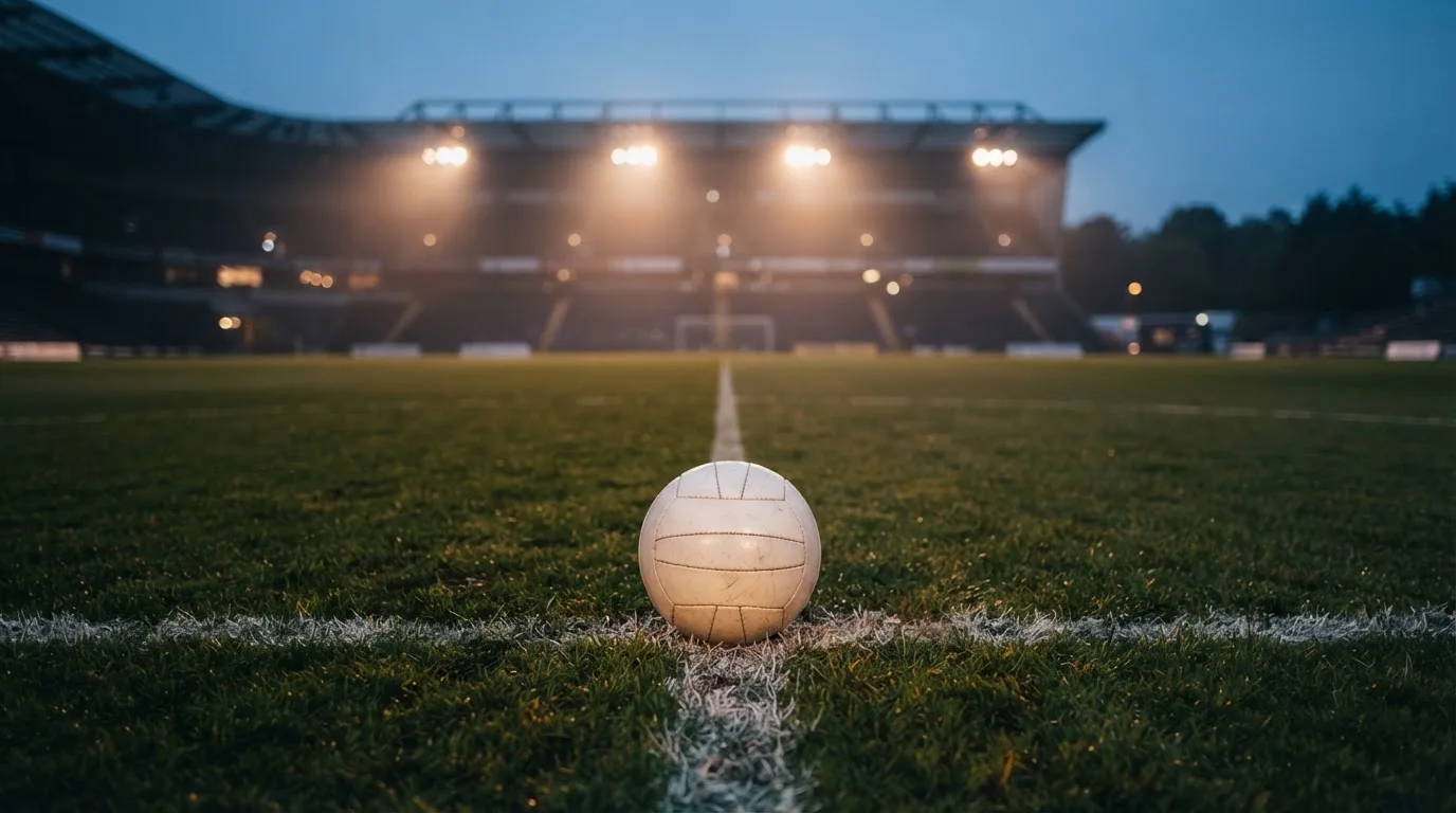 Ein Fußball liegt auf dem Anstoßpunkt eines leeren Rasenplatzes, im Hintergrund steht ein Stadion unter Flutlicht in der Dämmerung.