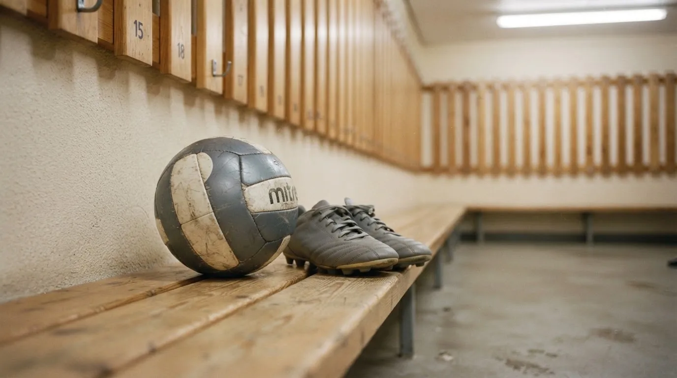 Ein Fußball liegt auf einer Holzbank in einer leeren Umkleide neben einem Paar Fußballschuhen ohne Markenzeichen.