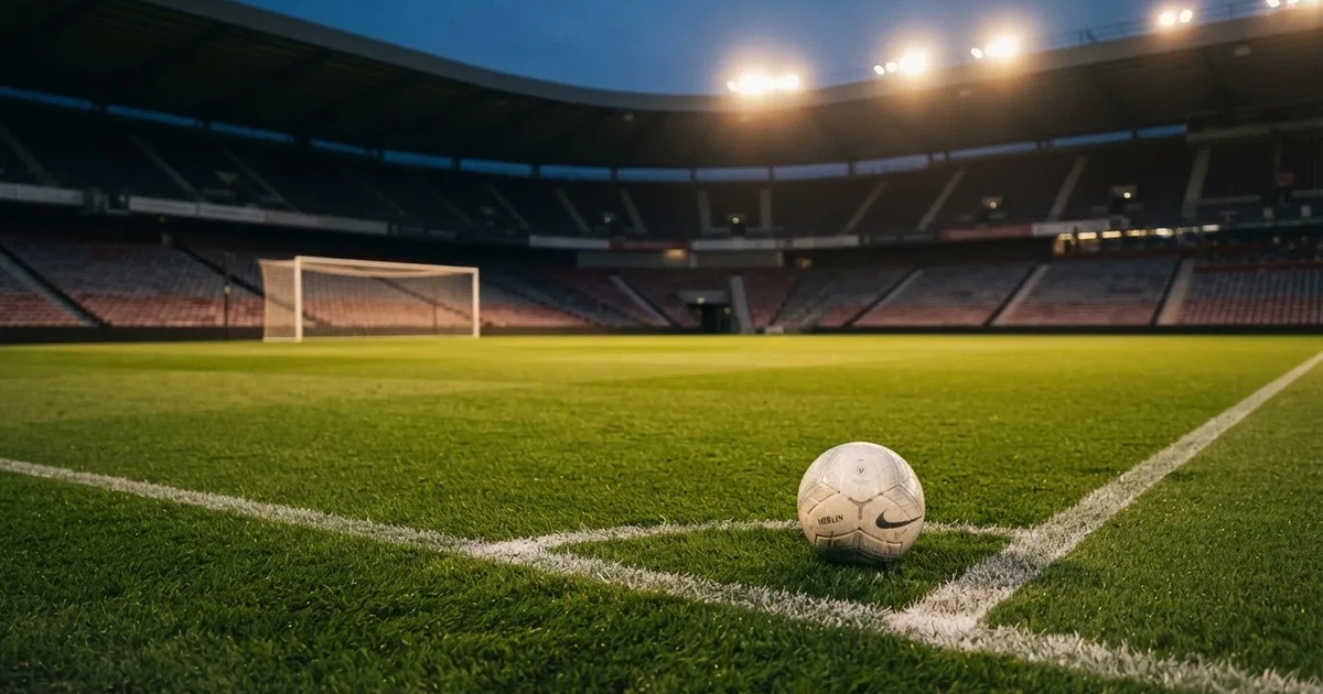 Ein Fußballplatz mit frisch gemähtem Rasen im Abendlicht, ein Ball liegt im Strafraum, im Hintergrund unscharf eine leere Tribüne.