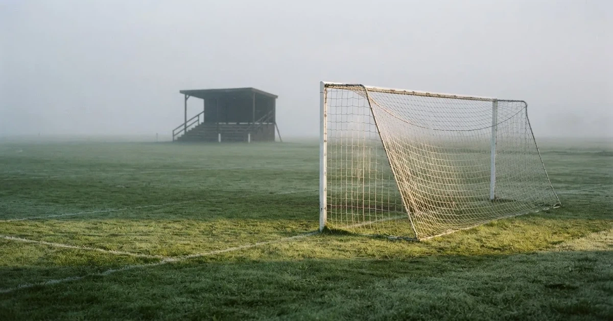 Ein Fußballtor mit Netz steht im Morgennebel auf einem leeren Rasenplatz, im Hintergrund die Silhouette einer kleinen Tribüne.