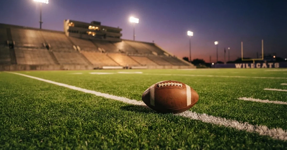 Ein American-Football-Ball liegt auf einem Rasenplatz unter Flutlicht, im Hintergrund eine große Tribüne in der Dämmerung.