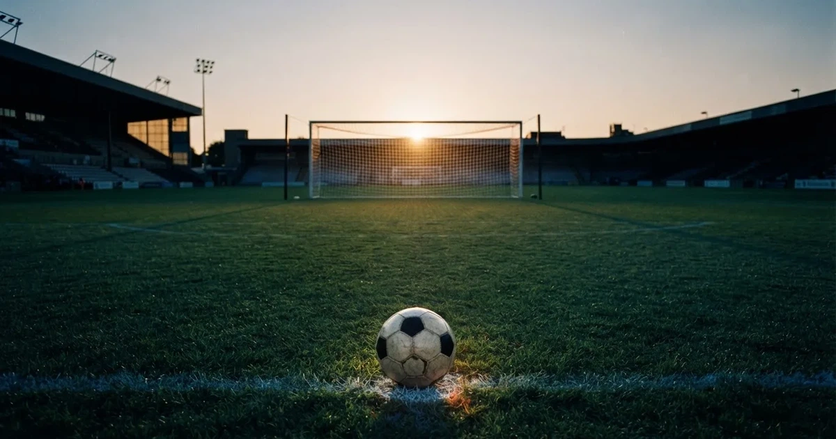 Ein Fußballtor mit Netz im Gegenlicht auf einem leeren Platz am Abend, davor ein Ball auf dem Rasen.