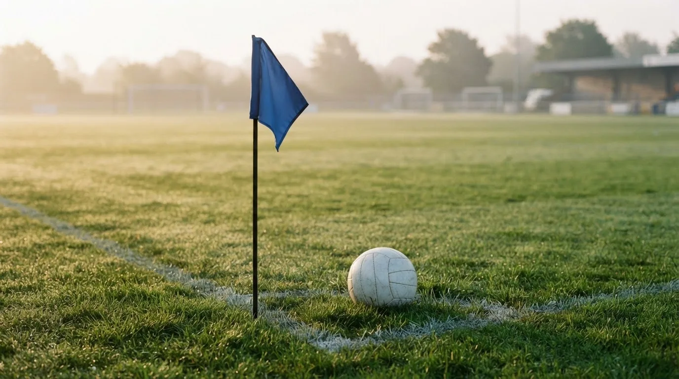 Ein Fußball liegt auf Rasen neben einer Eckfahne ohne Aufdruck auf einem leeren Platz im Morgenlicht.